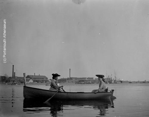 Two women rowing in Piscataqua River with Kittery, Maine shipyard (c) Portsmouth Athenaeum